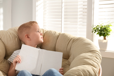 Photo of Cute little boy with book in papasan chair at home