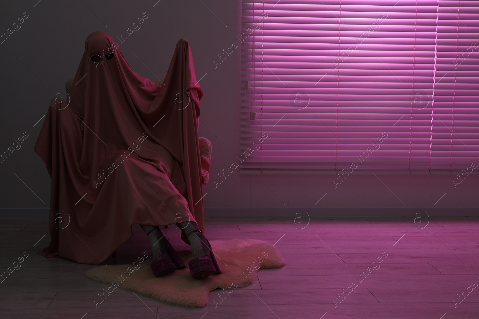 Photo of Glamorous ghost. Woman in sheet and high heel shoes on armchair in pink light, space for text