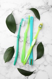 Photo of Colorful plastic toothbrushes and green leaves on white marble table, flat lay