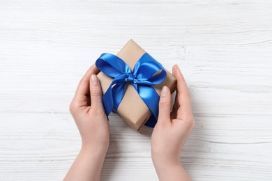 Woman holding gift box with blue bow at white wooden table, top view