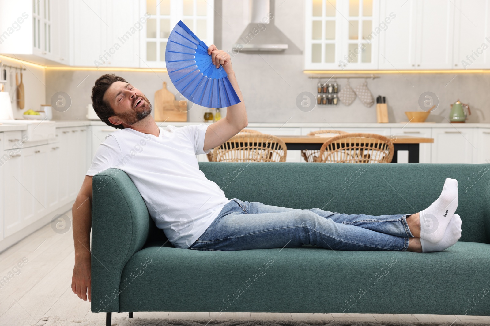 Photo of Bearded man waving blue hand fan to cool himself on sofa at home