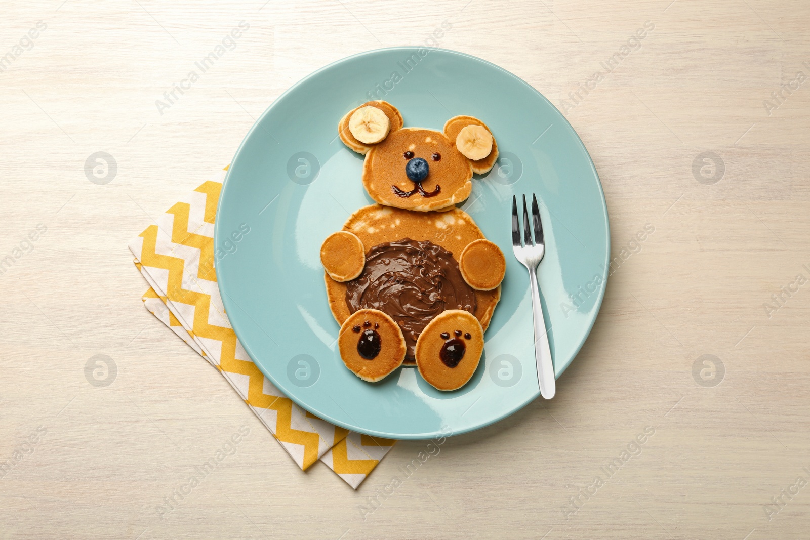 Photo of Creative serving for kids. Plate with cute bear made of pancakes, blueberries, bananas and chocolate paste on light wooden table, top view