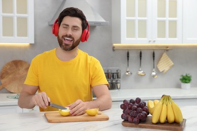 Photo of Handsome man listening music with headphones while cutting apple at white marble table in kitchen. Space for text