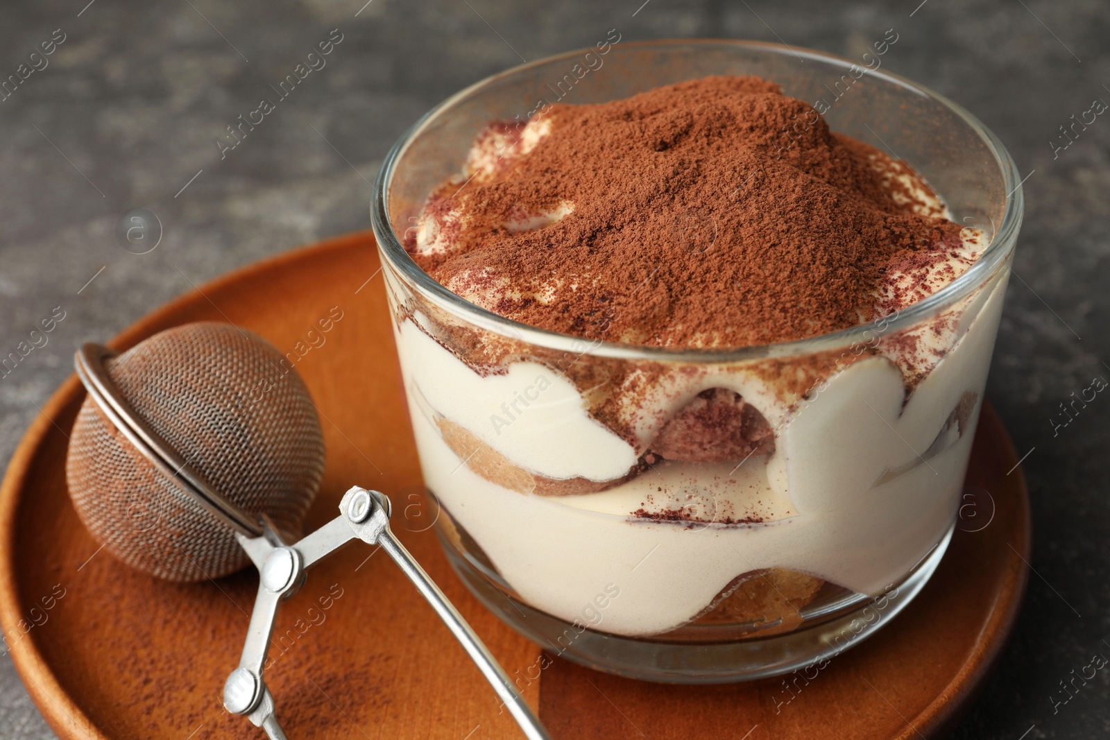 Photo of Delicious tiramisu in glass and sieve with cocoa powder on grey table, closeup