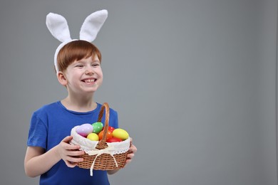 Easter celebration. Cute little boy with bunny ears and wicker basket full of painted eggs on grey background. Space for text