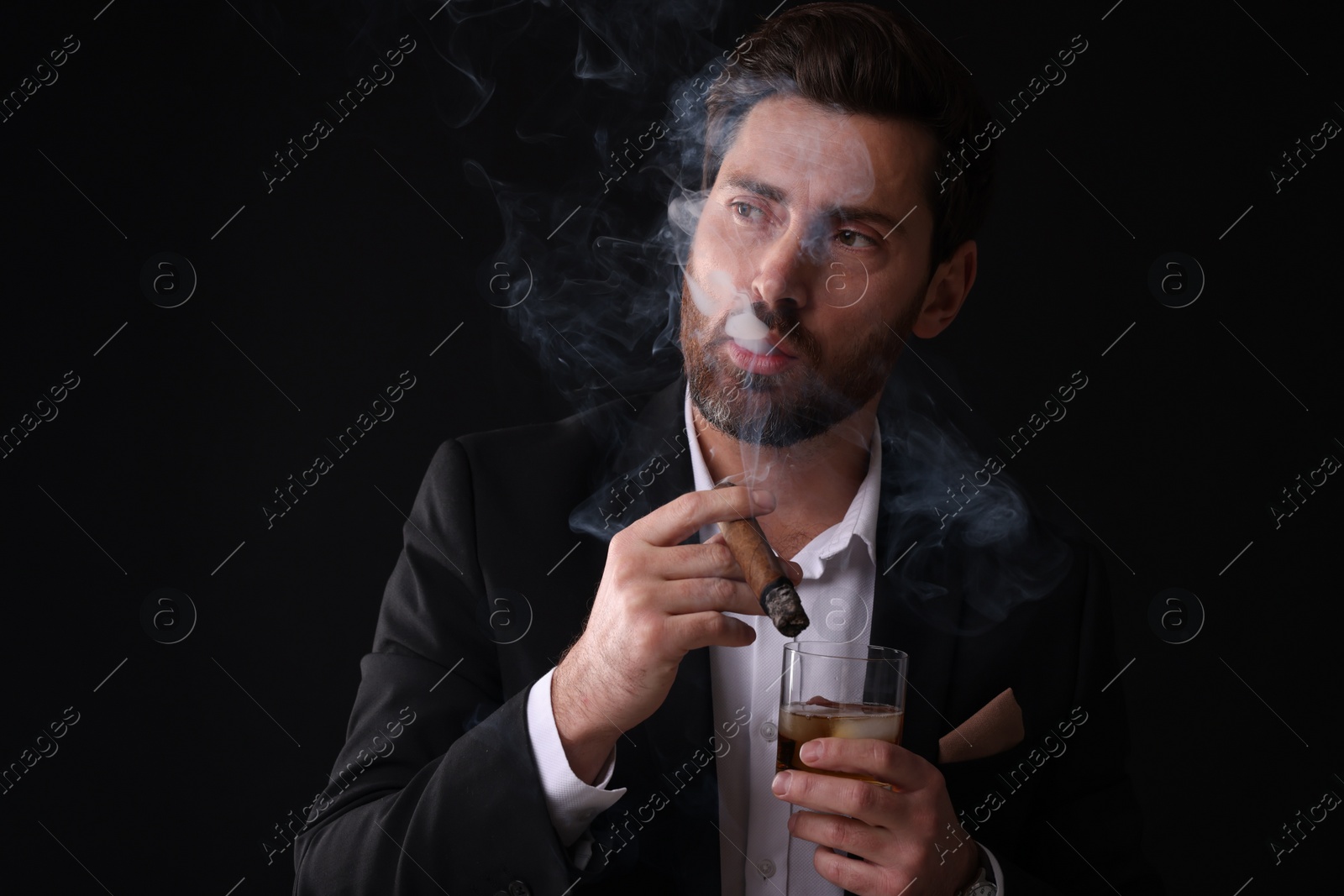 Photo of Handsome man in elegant suit with glass of whiskey smoking cigar on black background