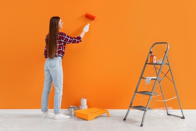 Photo of Designer painting orange wall with roller near folding ladder indoors