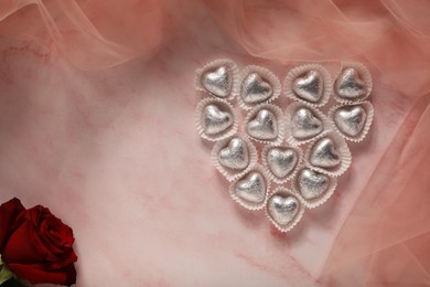 Heart made with delicious chocolate candies and beautiful rose on pink table, flat lay