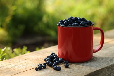 Photo of Red cup of bilberries on wooden table outdoors, space for text