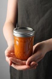 Photo of Woman holding glass jar of delicious persimmon jam on gray background, closeup