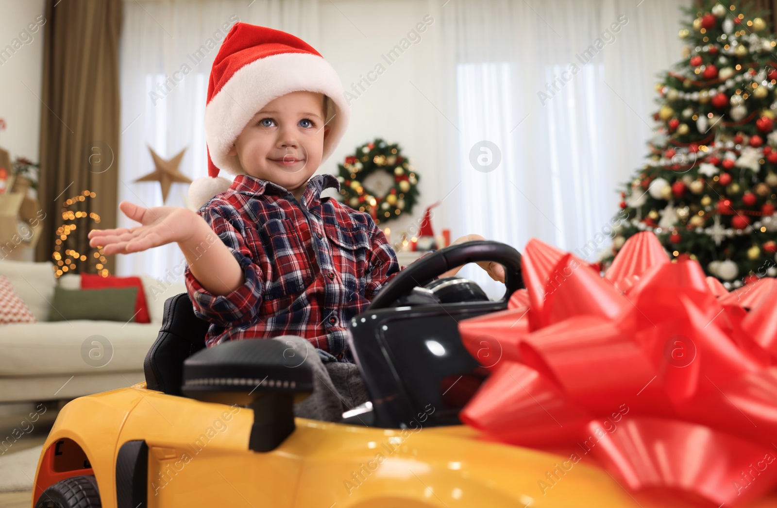 Photo of Cute little boy sitting inside toy car in room decorated for Christmas