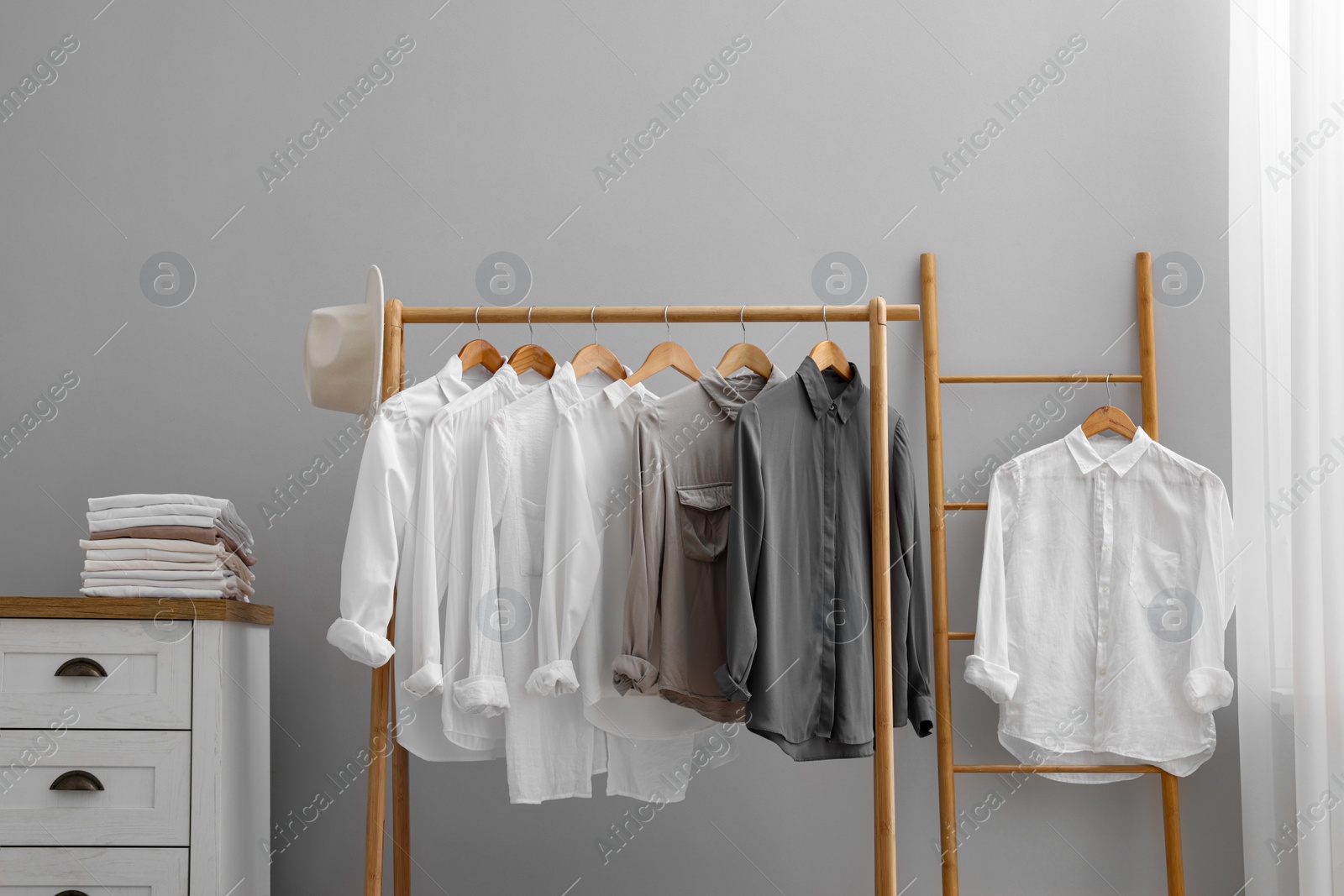 Photo of Rack with different stylish shirts, hat and chest of drawers near grey wall indoors. Organizing clothes