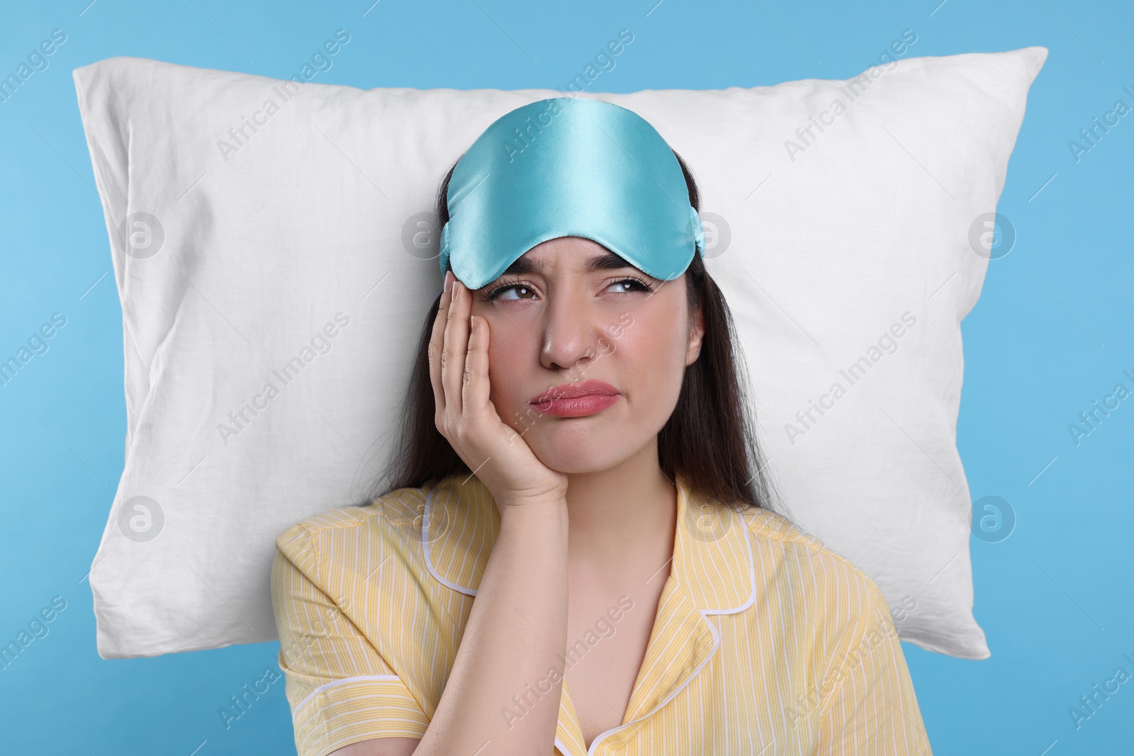 Photo of Tired young woman with sleep mask and pillow on light blue background. Insomnia problem