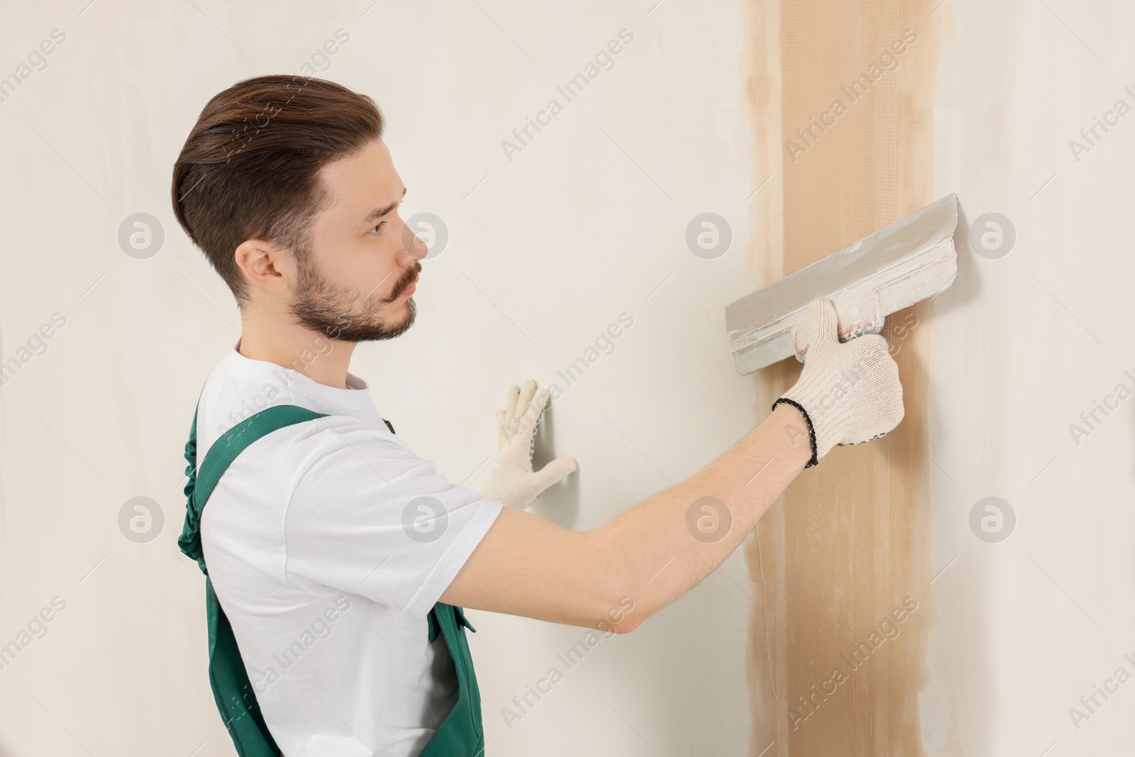 Photo of Worker in uniform plastering wall with putty knife indoors