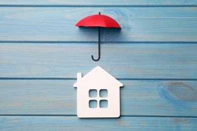 Small umbrella and house figure on light blue wooden background, flat lay