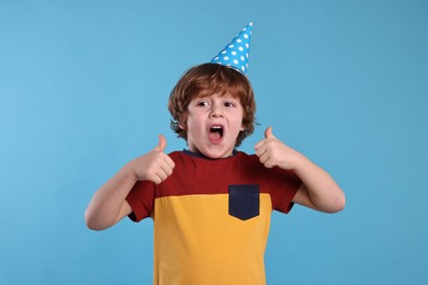 Birthday celebration. Cute little boy in party hat showing thumbs up on light blue background