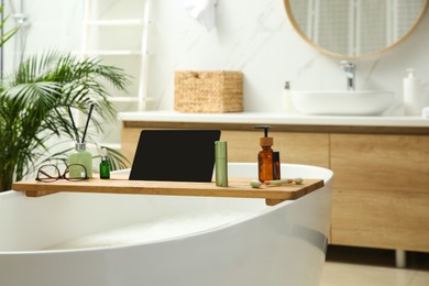 Photo of Wooden bath tray with tablet, face roller and cosmetic products on tub indoors