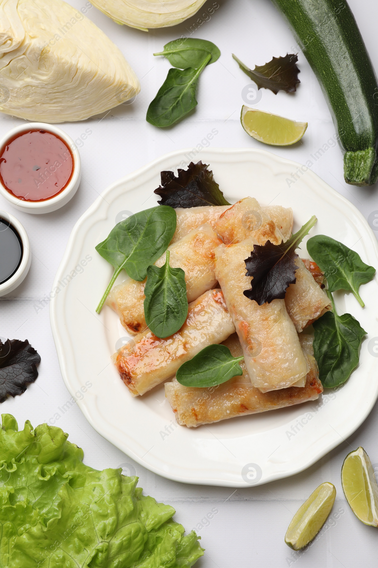 Photo of Plate with tasty fried spring rolls, spinach, sauces and other products on white tiled table, flat lay