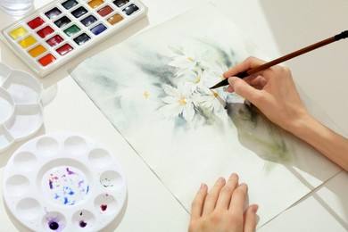 Photo of Woman painting flowers with watercolor at white table, above view. Creative artwork