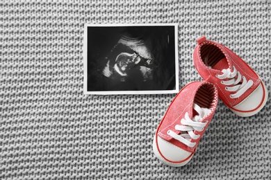 Photo of Ultrasound photo of baby and cute boots on knitted blanket, flat lay. Space for text