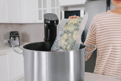 Photo of Woman putting vacuum packed broccoli into pot with sous vide cooker in kitchen, closeup. Thermal immersion circulator
