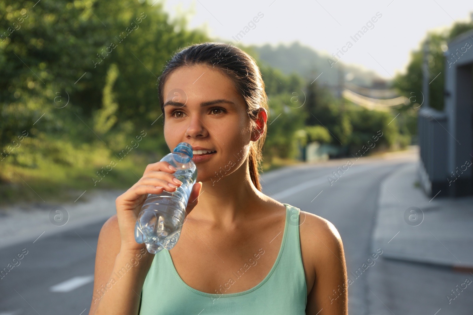 Photo of Happy young woman drinking water outdoors on hot summer day. Refreshing drink