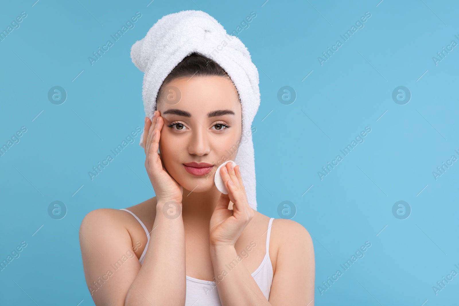 Photo of Beautiful woman in terry towel removing makeup with cotton pad on light blue background, space for text