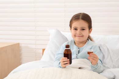 Photo of Effective medicine. Cute girl holding bottle and measuring cup with cough syrup in bed, space for text