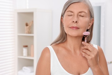 Woman massaging her face with rose quartz roller in bathroom. Space for text
