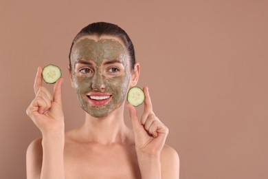 Photo of Beautiful woman with clay mask holding pieces of cucumber on light brown background, space for text