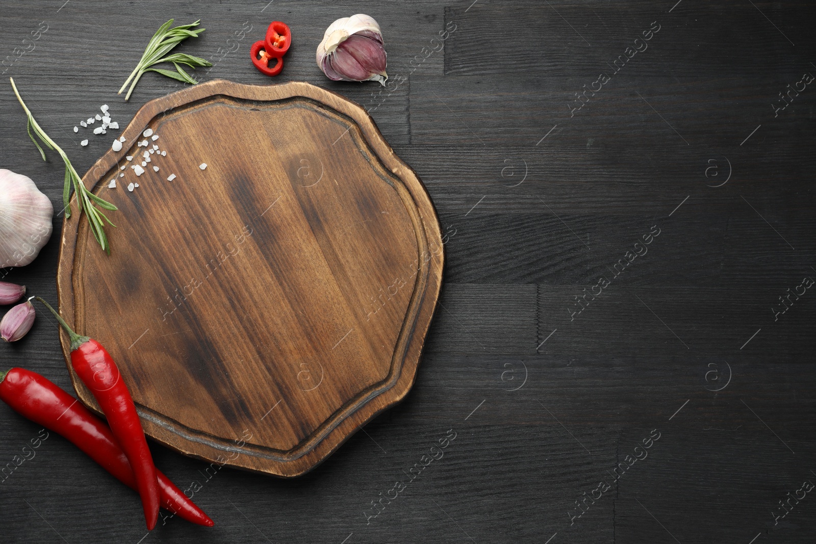 Photo of Cutting board, garlic, rosemary and chili peppers on black wooden table, flat lay. Space for text