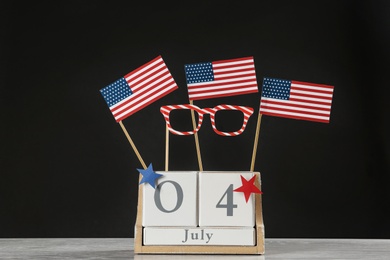 Photo of Wooden calendar with USA flags and paper glasses on table against black background. Happy Independence Day