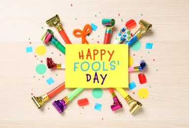 Card with text Happy Fool's Day and party blowers on wooden background, flat lay 