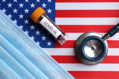 Photo of Test tube with blood sample, medical mask and stethoscope on American flag, flat lay. Coronavirus pandemic in USA