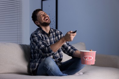 Laughing man with popcorn bucket watching comedy via TV at home in evening