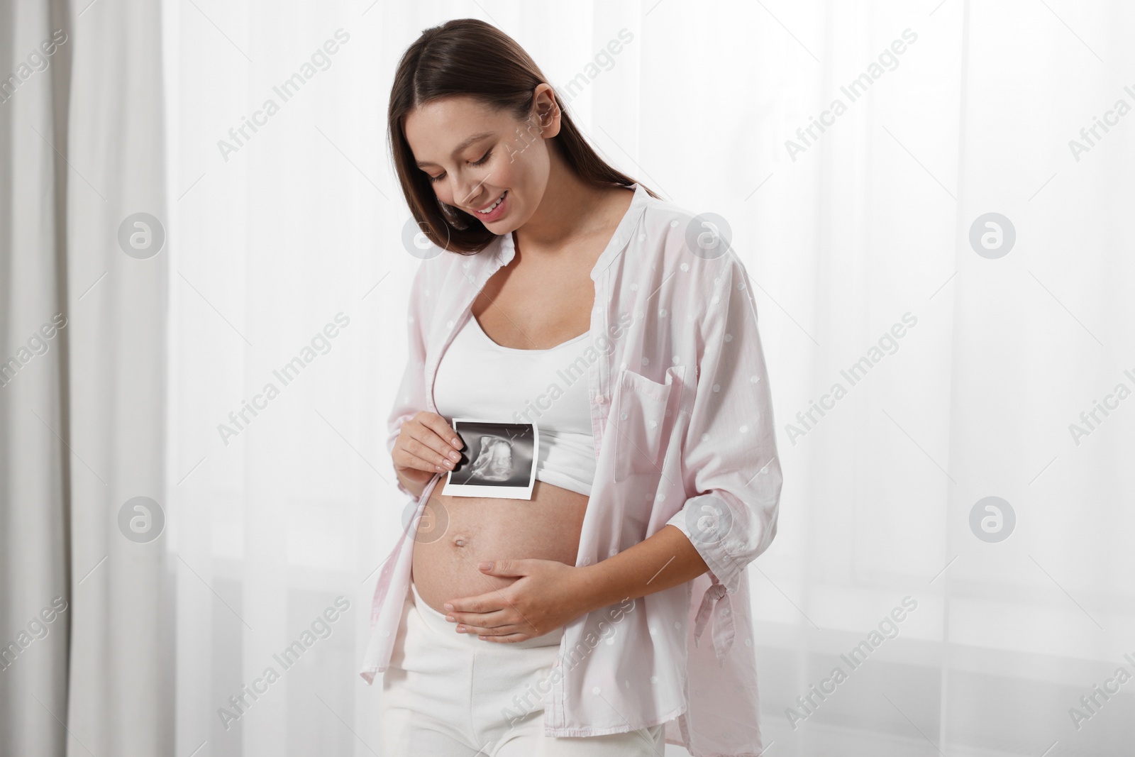 Photo of Beautiful pregnant woman with ultrasound picture of baby near window indoors