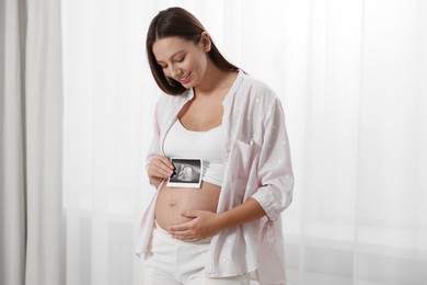 Photo of Beautiful pregnant woman with ultrasound picture of baby near window indoors