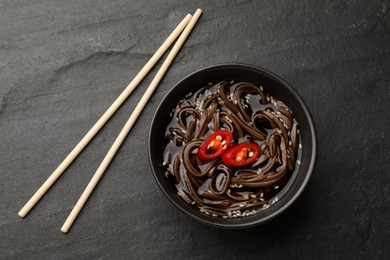 Photo of Tasty soup with buckwheat noodles (soba), chili pepper in bowl and chopsticks on dark grey textured table, flat lay