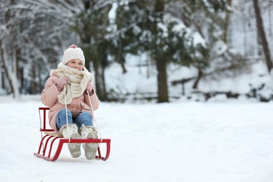 Photo of Cute little girl enjoying sledge ride through snow in winter park, space for text
