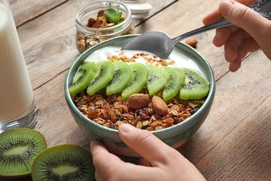 Image of Woman eating tasty granola with yogurt and sliced kiwi for breakfast at wooden table, closeup