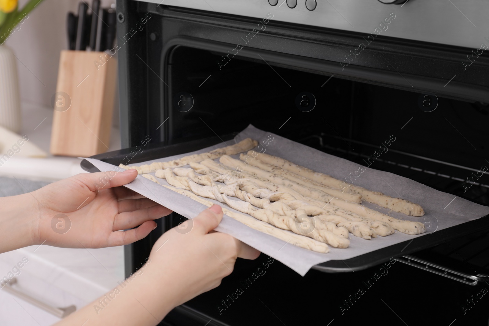 Photo of Woman putting baking sheet with homemade breadsticks into oven in kitchen, closeup. Cooking traditional grissini