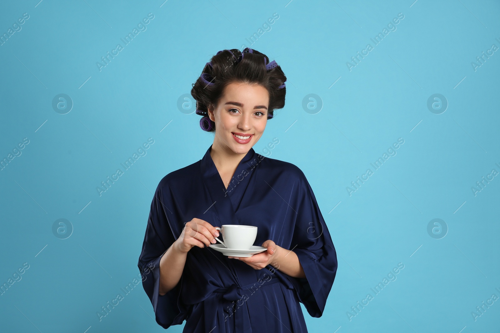 Photo of Happy young woman in silk bathrobe with hair curlers holding cup of drink on light blue background