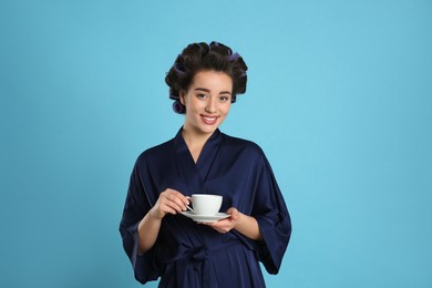 Happy young woman in silk bathrobe with hair curlers holding cup of drink on light blue background