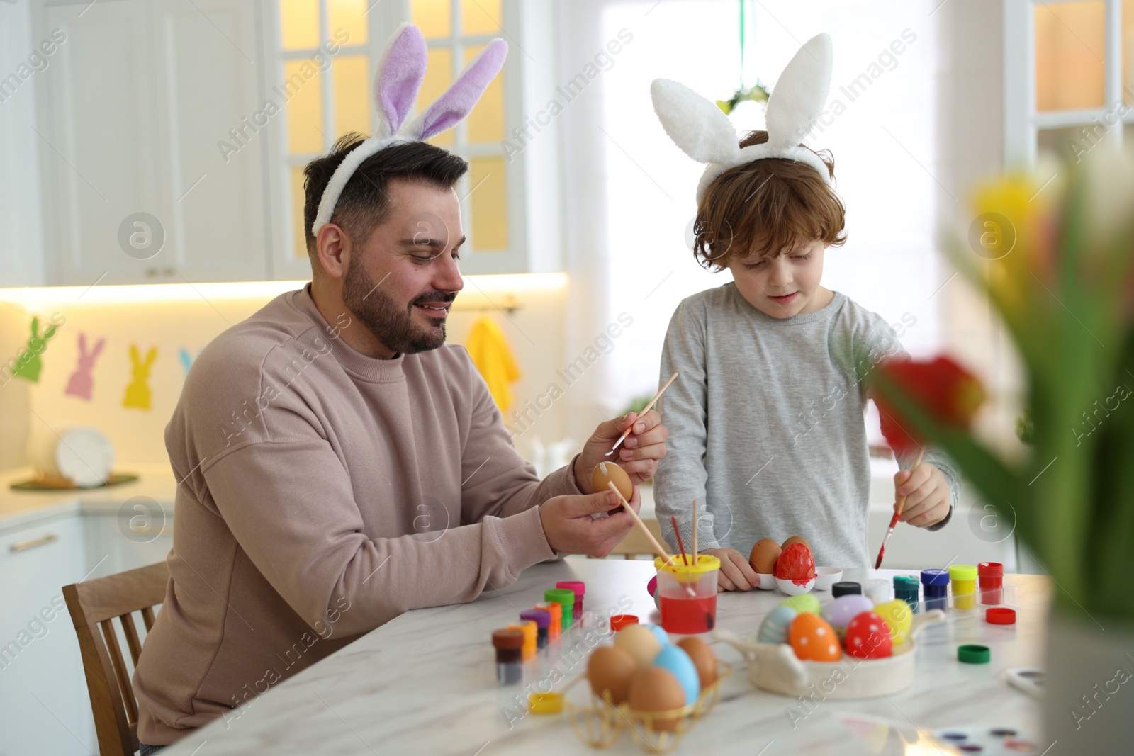 Photo of Easter celebration. Father with his little son painting eggs at white marble table in kitchen