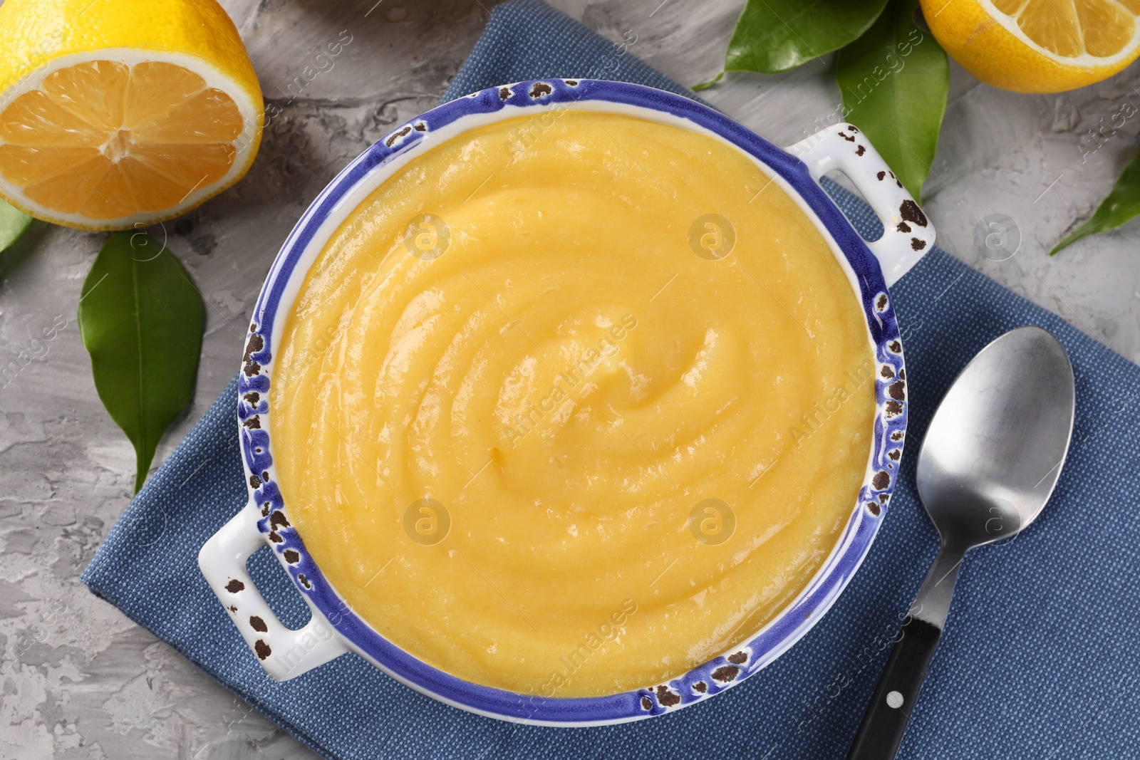 Photo of Delicious lemon curd in bowl, fresh citrus fruit, green leaves and spoon on grey table, top view
