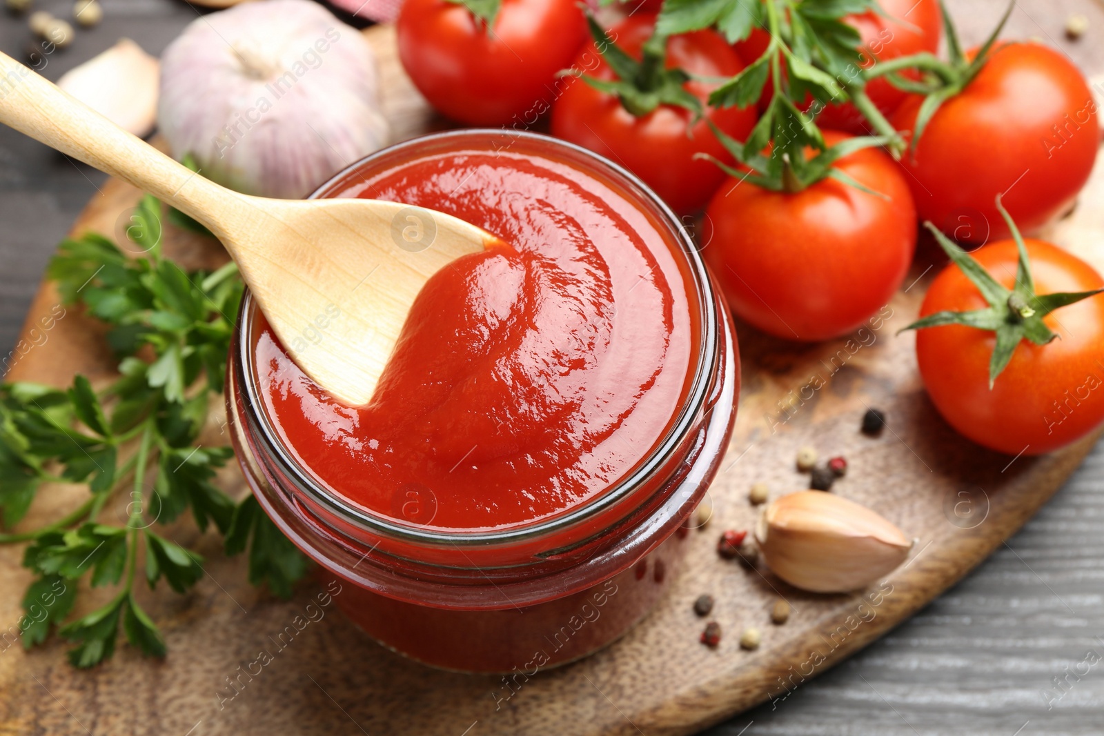 Photo of Jar and spoon with tasty ketchup, fresh tomatoes, parsley and spices on grey wooden table, closeup