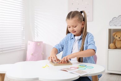 Photo of Motor skills development. Smiling girl playing with geoboard and rubber bands at white table in room. Space for text