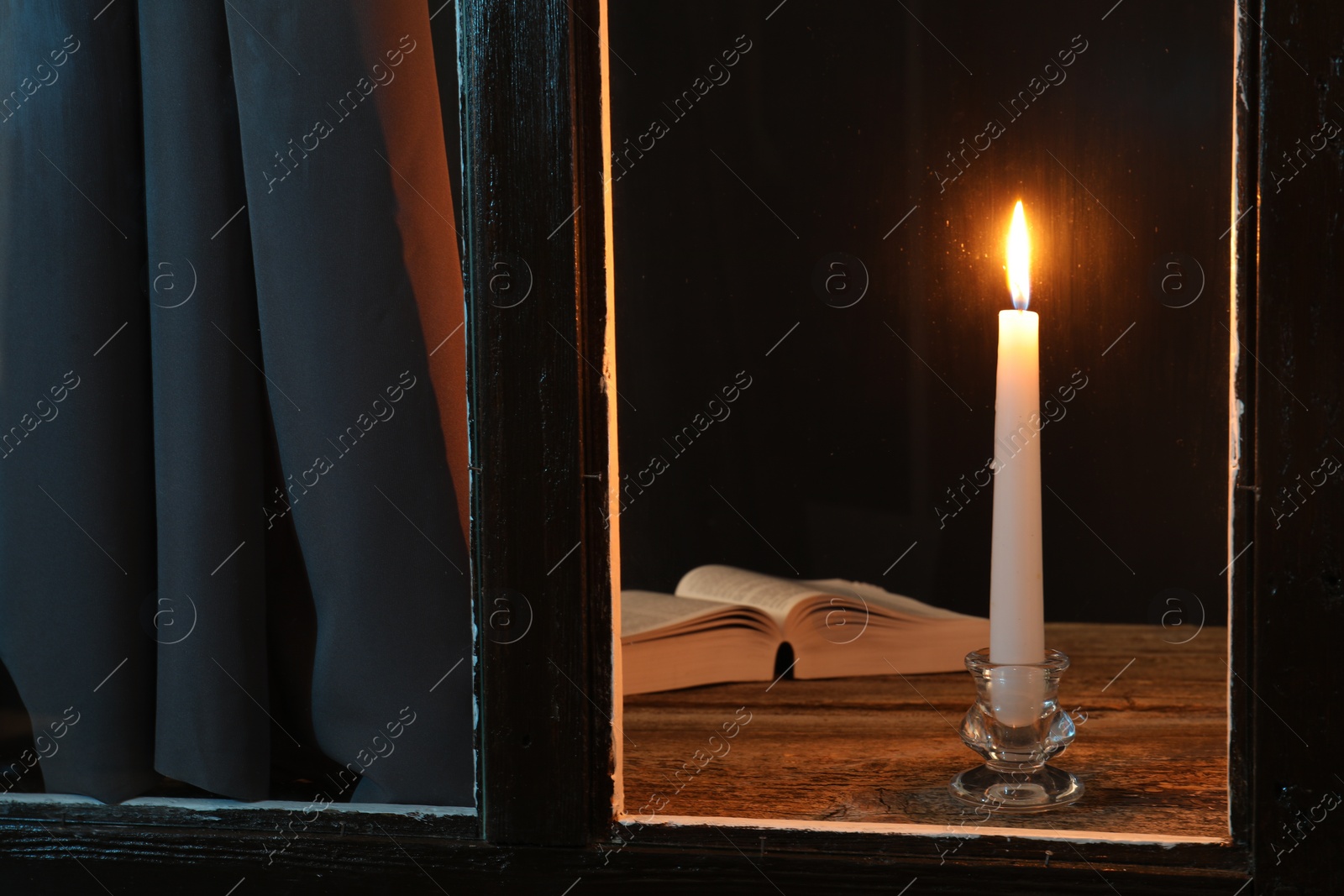 Photo of Burning candle and Bible on wooden table at night, view through window