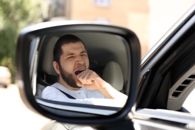 Photo of Tired man yawning in his modern car, view through car side mirror