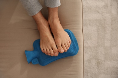Photo of Man warming feet with hot water bottle on sofa, closeup